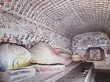 Photo couleur de peintures murales bouddhiques à l'intérieur d'une grotte. À gauche : une statue de bouddha allongé sur un socle en pierre, avec, le long du mur, des statuettes représentant divers personnages.