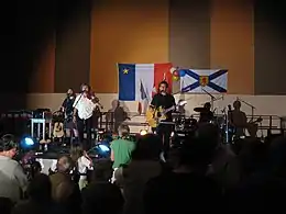 Photographie du groupe de musique Suroît durant l'un de ses spectacles. Sur le mur au fond de la scène se trouvent le drapeau de l'Acadie et celui de la Nouvelle-Écosse.