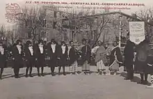 Carte postale noir et blanc d'un groupe d'une douzaine de personnes debout. Un bâtiment an arrière-plan.