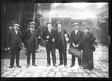 Groupe de cinq hommes et une femme, portant chacun un sac de voyage, une musette ou un paquet, une casquette ou un chapeau, certains en cravate, tous en veste.