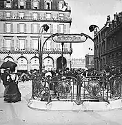 Photo noir et blanc d'une place avec une balustrade et de hauts candélabres autour d'un escalier ; devant à gauche, une femme en jupe longue ; derrière, un grand immeuble