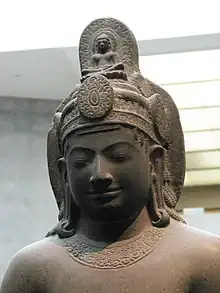 Sur la statue en grès le Bodhisattva a les yeux clos et a une coiffe au sommet de laquelle trône une représentation de bouddha