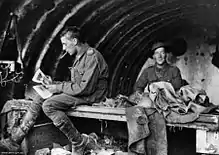 Deux hommes en uniforme militaire dans un bâtiment. L'homme de gauche a quelque chose dans la bouche et est assis en train de regarder des papiers, en s'éloignant de la caméra. L'homme de droite sourit et regarde la caméra. Le toit de la structure est incurvé.