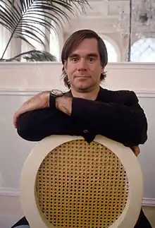 A man wearing a long-sleeved black shirt and watch; his arms and crossed, resting on the back of a chair.