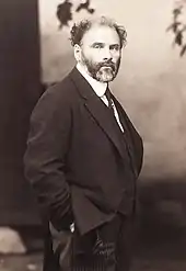 Photographie en noir et blanc de Gustave Klimt, barbu, les mains dans les poches de son pantalon.