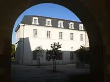 Photo prise sous un proche dont l'arrondi sombre coupe le ciel bleu ; on voit un bâtiment de deux étages aux murs blancs, le toit mansardé est fait d'ardoises.