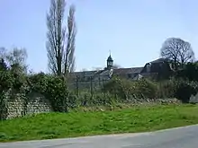 Derrière une végétation sauvage, on aperçoit la lanterne de la chapelle, les étages du bâtiment principal et, à droite, le pavillon nord. Au premier plan, brèche dans le mur d'enceinte, que colmate un grillage en mauvais état.