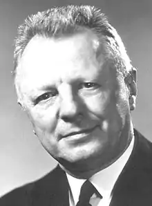 Head and shoulder of a 60-ish man with a flattop haircut and in a coat and tie, looking directly at camera with head tilted to his right and a slight smile.