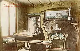 Photo sépia colorisée par endroits montrant un homme barbichu à un bureau dans un décor chargé