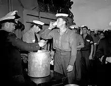 Photographie d'époque du Splice the mainbrace célébré le lendemain de la reddition officielle du Japon à bord d'un navire de guerre canadien. Les marins se tiennent en ligne devant un bar improvisé sur le pont où l'on sert du rhum. Un officier note les noms sur un calepin.