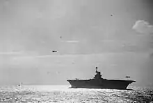 Photo en noir et blanc d'un porte-avions vu de bâbord sur lequel un avion apponte, pendant qu'un autre patrouille au-dessus.