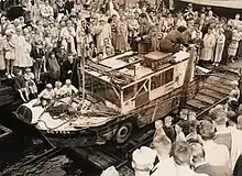 Photographie noir et blanc d'une foule autour d'une jeep amphibie modifiée. Une femme, sur le toit, sert des mains.