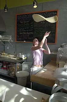 Derrière le comptoir, une jeune femme fait sauter une pâte de pizza, qui tournoie en l'air au-dessus de sa tête. Elle regarde sa pâte et ses mains sont prêtes à la rattraper. Devant elle, le plan de travail en bois est légèrement fariné.