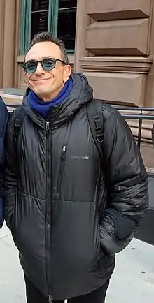 Photographie de plein pied d'un homme souriant qui porte des lunettes de soleil et une doudone noire.