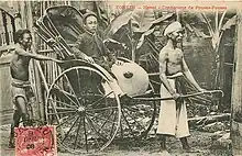 Un homme asiatique en habit traditionnel, assis dans un pousse-pousse tiré par son conducteur, ce dernier étant torse nu. Un autre homme, vêtu d'un pagne, se tient debout derrière le véhicule.