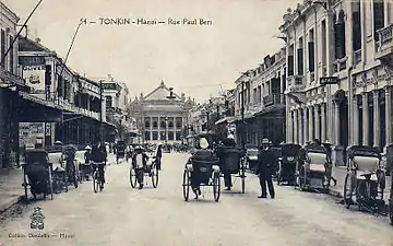 Photo ancienne montrant une rue dans laquelle circulent des pousse-pousses, avec en arrière-plan un grand bâtiment.