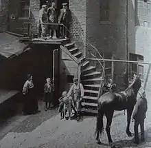 Photo en noir et blanc d'une cour d'immeuble, un cheval avec son propriétaire se trouvant au bas à droite de la photo, une femme et des enfants se tenant dans la cour, et trois hommes se tenant en haut des marches d'escalier.