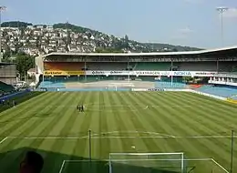 Vue d'un stade de football aux tribunes vides avec une colline et des maisons en arrière-plan