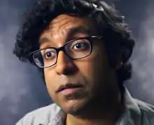 Photographie du visage d'un homme. Il porte des lunettes et à des cheveux mi-longs noirs.