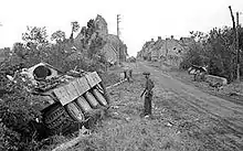 photo montrant un soldat anglais devant la carcasse d'un char allemand à l'entrée d'un village