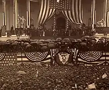 Photographie d'une estrade décorée par des drapeaux américains. De nombreux participants tiennent des parapluies et toutes les personnes de la foule en contrebas portent des chapeaux.