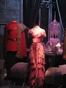 Deux mannequins : celui de gauche porte un costume rouge avec une cape en fourrure sur son épaule gauche et celui de droite porte une longue robe rose à volants