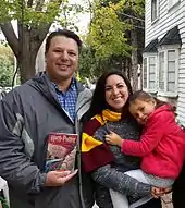 Un homme (à gauche) et une femme (à droite) avec une écharpe rouge et or et tenant une fillette dans ses bras (l'homme montre un livre Harry Potter au photographe et les trois personnes sourient)