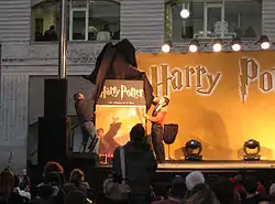 Deux hommes retirant un drap noir d'une couverture de livre géante, dans les tons orange et noir, montrant Harry la main levée et lançant un sortilège