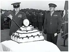 Devant un groupe d'hommes, un homme coupe un gâteau avec une épée alors qu'un second homme le regarde.
