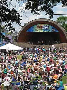 Scène de concert en plein air sous une arche avec public dans le gazon au premier plan.