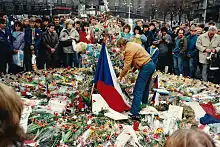 Un homme de profil au milieu d'une foule en recueillement, aux côtés d'un drapeau tchécoslovaque, le sol est tapissé de roses.