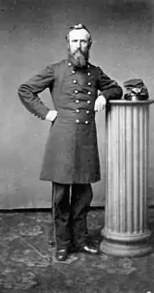 Un homme barbu portant un uniforme militaire avec un long manteau à boutons est accoudé à un pilier où est posé un képi.