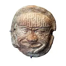 Pièce d'argile plate et ronde sur laquelle est représentée une tête humanoïde aux yeux graves et au rictus prononcé.