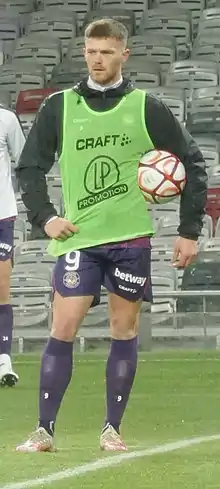 Rhys Healey en tenue d'échauffement, short violet et imperméable noir, de face, sur la pelouse du Stadium