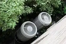 Deux cylindres gris sortant du sol avec des trous pour laisser passer de l'air et connectés à l'intérieur d'un bâtiment par des tuyaux noirs.