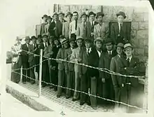 Photo d'archive en noir et blanc de plusieurs hommes habillés à l'occidentale derrière une clôture.