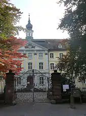 Bâtiment baroque avec un fronton et un clocheton, peint en jaune clair derrière une grille de fer forgé.