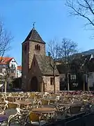 Photo couleur d'une tour d'église adossée à un bâtiment, sur une place encombrée de tables et de chaises.