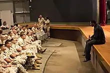 Un homme, assis sur une estrade, micro en main, se tient devant une assemblée de militaires en tenue.
