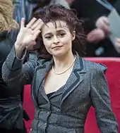 Une femme brune d'une quarantaine d'années, aux cheveux longs et attachés, faisant un signe de sa main droite au photographe.