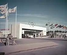 Une grande arche rectangulaire ornée des anneaux olympiques. À gauche, des drapeaux olympiques ; à droite de l'arche, un drapeau par pays. Une voiture est stationnée sous l'arche. La photo est en couleur.