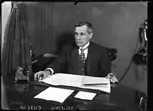 Photographie en noir et blanc d'un homme en costume assis à son bureau.