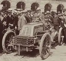 Photo d'un homme en tenue de ville prenant la pose au volant de son véhicule devant un parterre de spectateurs.