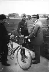 Photographie en noir et blanc montrant un cycliste debout à côté de son vélo, entouré de deux hommes.