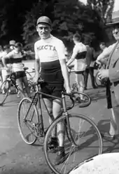 Photographie en noir et blanc d'un coureur debout à côte de son vélo portant un maillot marqué Dilecta.