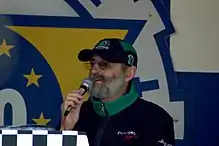 photo d'un homme, habillé en noir et vert, parlant dans un micro, de face, trois-quart
