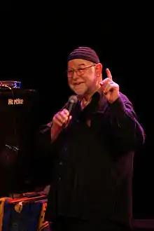 un homme avec des lunettes, une barbe blanche et un petit bonnet parle dans un micro.