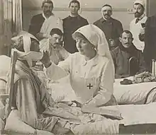 revêtue d'une tenue d'infirmière avec brassard et coiffe portant une croix rouge, Henriette, assise au chevet d'un blessé au visage bandé, tend la main vers ce soldat. À l'arrière-plan figurent cinq blessés et un médecin revêtu d'une blouse blanche et coiffé d'un calot clair