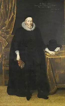 Homme âge portant une barbe, dans une longue robe noire à col gris.