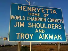 Un panneau bleu avec dessus les inscriptions en lettres blanches suivantes : « Henryetta, Home of World Champion Cowboys Jim Shoulders and Troy Aikman ».